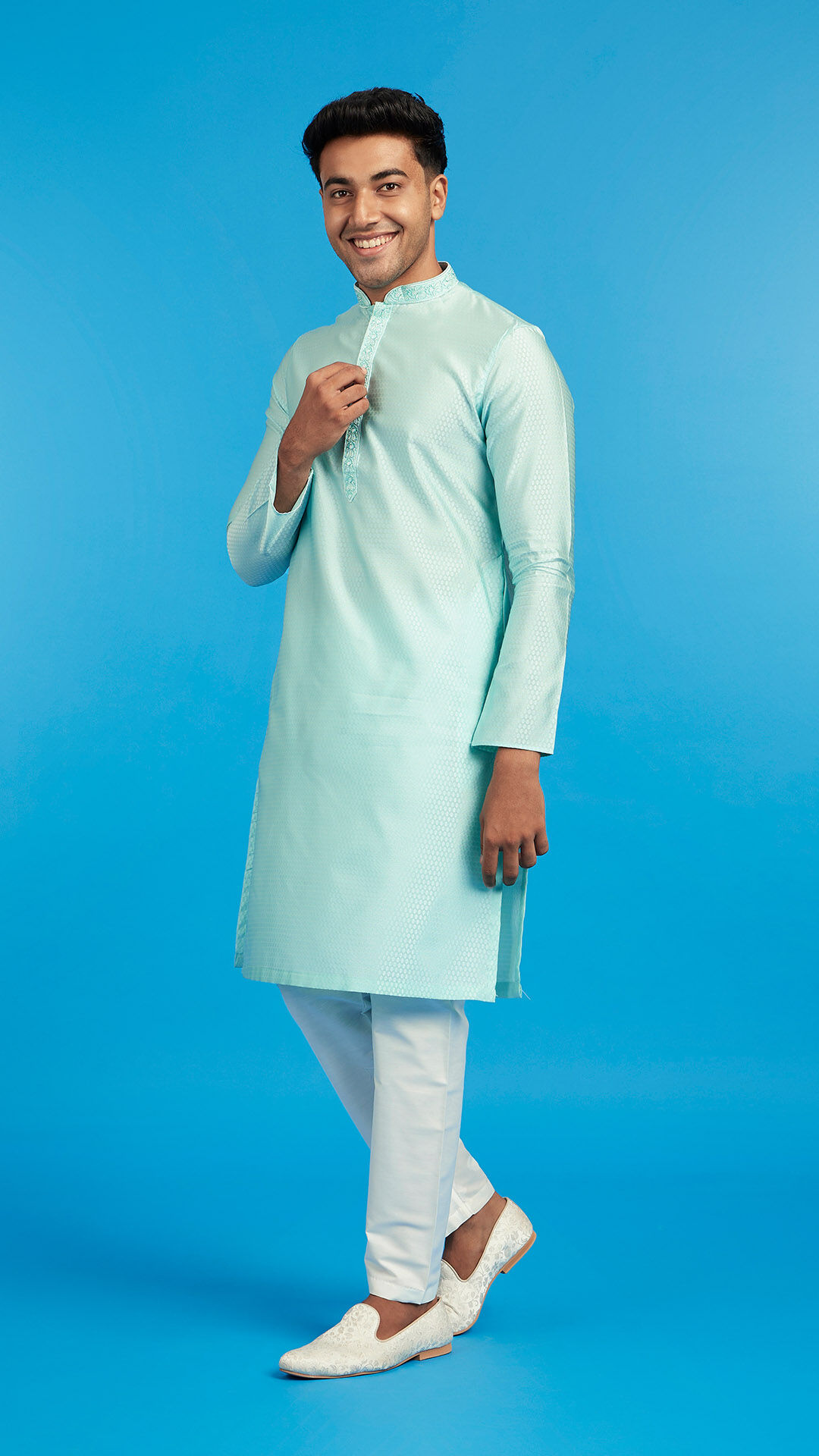 Smiling young Indian man in a pastel blue kurta with white churidar and embroidered loafers, posing against a blue background.